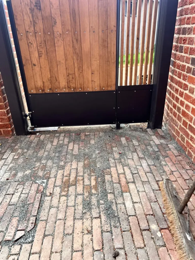 An open timber pedestrian gate in a brick surround on a brick-paved path